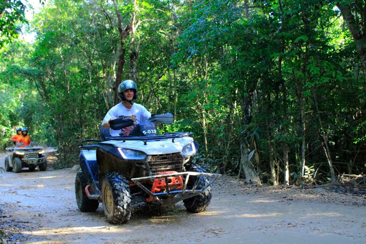 Hombre con casco conduciendo un ATV por un camino de terracería en medio de la selva, con otro conductor detrás.