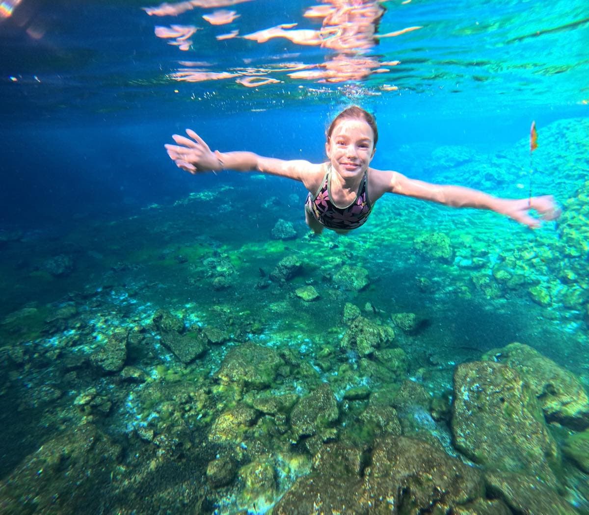 Niña nadando bajo el agua con los brazos abiertos en un cenote de agua turquesa, sobre un fondo de roca.