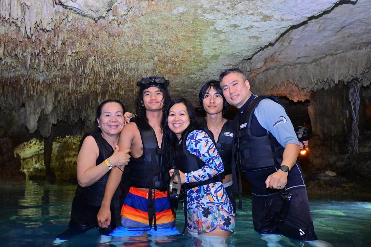 Familia de cinco personas con chalecos salvavidas, de pie en el agua de un cenote subterráneo con el techo cubierto de estalactitas.