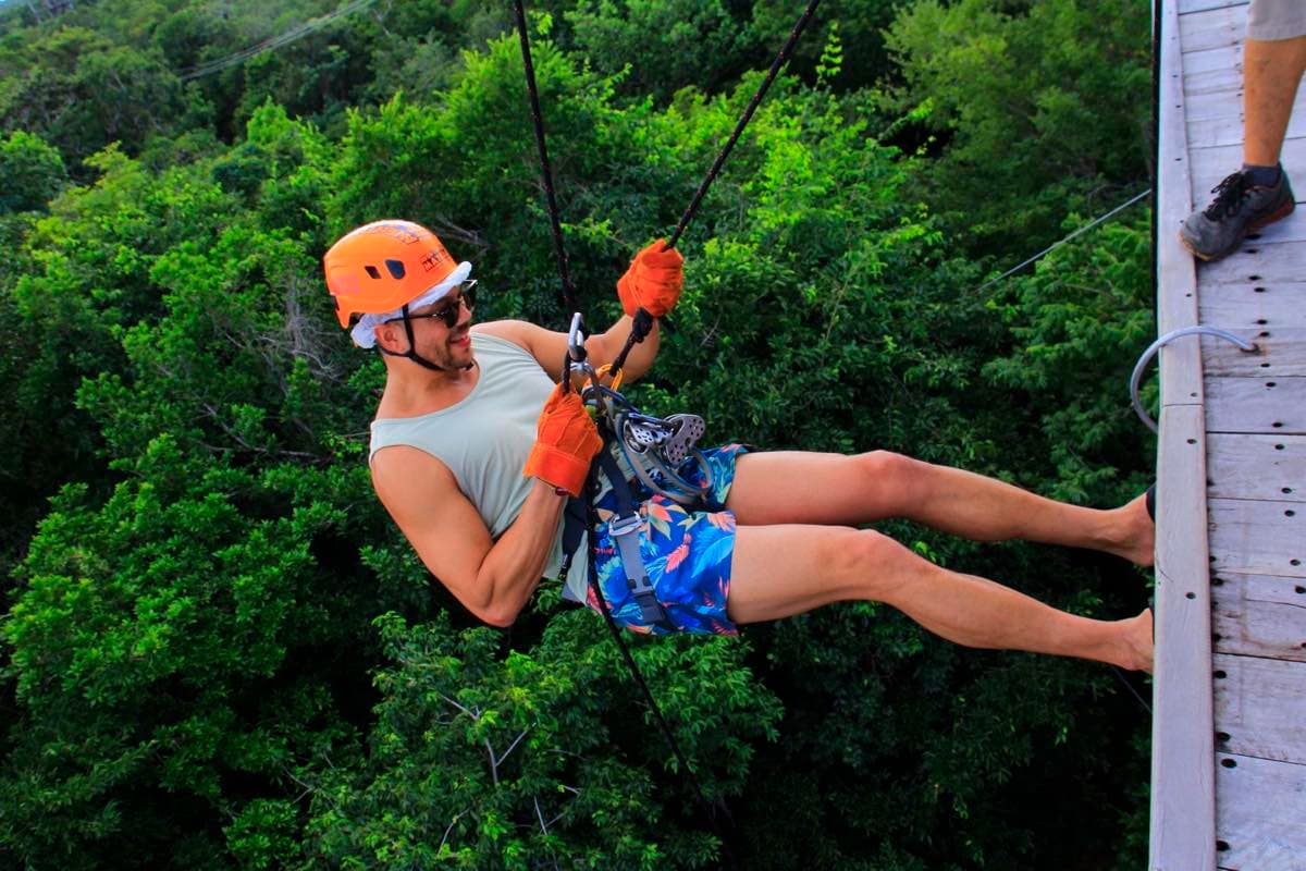 Hombre con casco naranja y guantes bajando en rapel desde una plataforma de madera, colgado sobre la copa de los árboles.